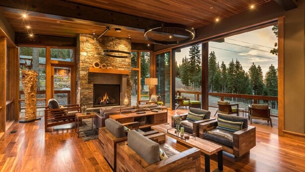 A spacious living room with a stone fireplace, wooden furniture, and large windows offering a view of the forest. - Modern Ski In Out Home Mid-mountain Northstar (Truckee)