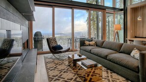 A modern living room with a large window, a fireplace, a sofa, and a coffee table. - Modern Ski In Out Home Mid-mountain Northstar (Truckee)
