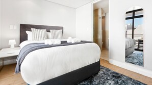 A modern hotel room with a large bed, a bedside table with a lamp, a mirror, and a view of the outside.