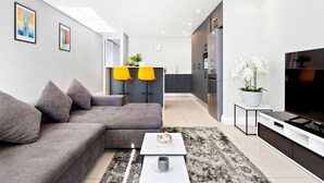 A modern living room with a grey sectional sofa, a coffee table, and a flat-screen TV. There is a kitchen area with a clock and a plant.