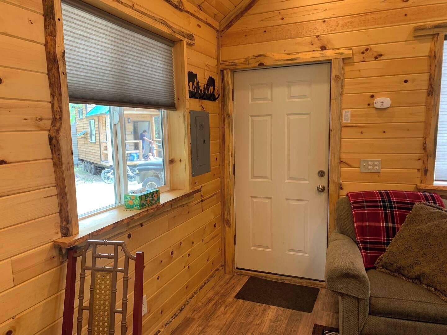Interior de una cabaña de madera con una puerta, una ventana con persianas, una pequeña mesa con una caja y un sofá con una manta.