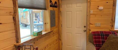 Interior de una cabaña de madera con una puerta, una ventana con persianas, una pequeña mesa con una caja y un sofá con una manta.