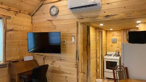 Interior de una cabaña de madera con una televisión montada en la pared, un reloj, un aire acondicionado, una mesa de comedor con sillas y una zona de cocina con estufa y refrigerador.
