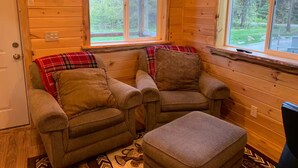 Interior acogedor de una cabaña con dos sillones, un pequeño reposapiés, paredes de madera, una ventana con vistas a la vegetación y una puerta.