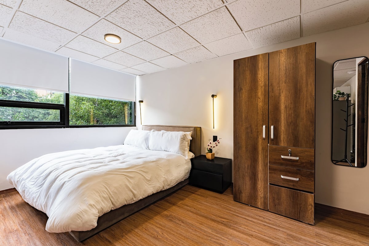 Un dormitorio con una cama grande, armario de madera, mesita de noche y ventana con vistas a la vegetación.