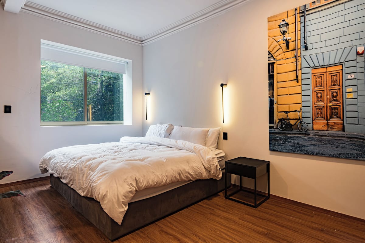 Un dormitorio con una cama grande, una mesita de noche pequeña, una ventana con vistas a la vegetación y una obra de arte colgada en la pared que muestra una escena callejera.