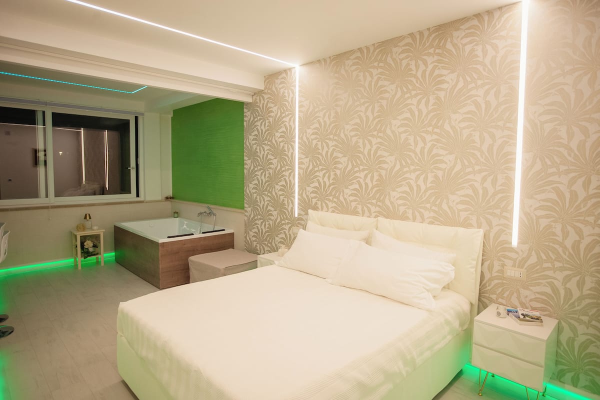A modern bedroom with a large bed, a small bedside table, a nightstand, a green accent wall, and a window with curtains.