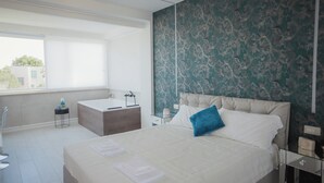 A modern hotel room with a large bed, a small desk, a window with blinds, and a bathroom with a bathtub. - B&B LA PERLA NERA (Fasano)