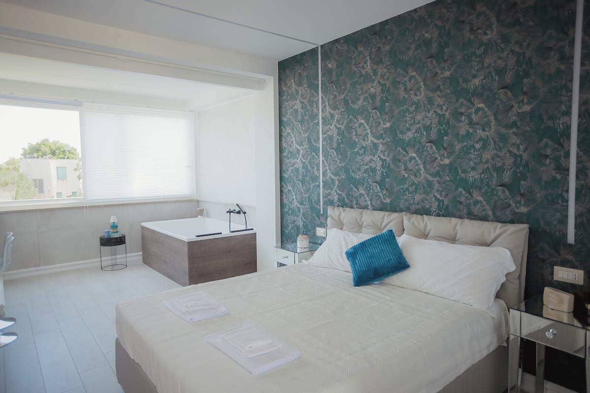 A modern hotel room with a large bed, a small desk, a window with blinds, and a bathroom with a bathtub.