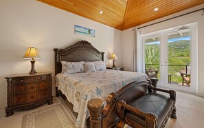 Quarto com cama grande, duas mesas de cabeceira com luminárias, cabeceira de madeira, vista para o exterior por uma porta de correr e um tapete decorativo.