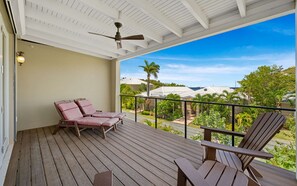A covered outdoor deck with wooden flooring, two lounge chairs, a ceiling fan, and a view of houses and greenery. - The Pirate's Landing North - St. John USVI - Chocolate Hole (Cruz Bay)