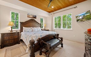 A bedroom with a large bed, wooden headboard, two bedside tables with lamps, a wooden bench, and a large window with a view of greenery.