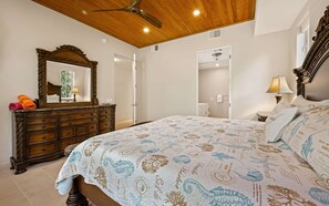 A bedroom with a wooden ceiling, a large bed with patterned bedding, a dresser with a mirror, and a ceiling fan. - The Pirate's Landing North - St. John USVI - Chocolate Hole (Cruz Bay)