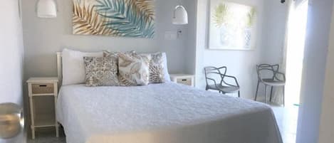 Un dormitorio con una cama grande, dos mesitas de noche, una mesita auxiliar y una silla. Hay dos lámparas de pared y dos obras de arte enmarcadas sobre la cama.
