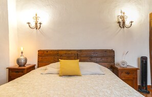 A bedroom with a wooden headboard, two wall-mounted lamps, a bedside table with a lamp, a radio, and a standing heater.