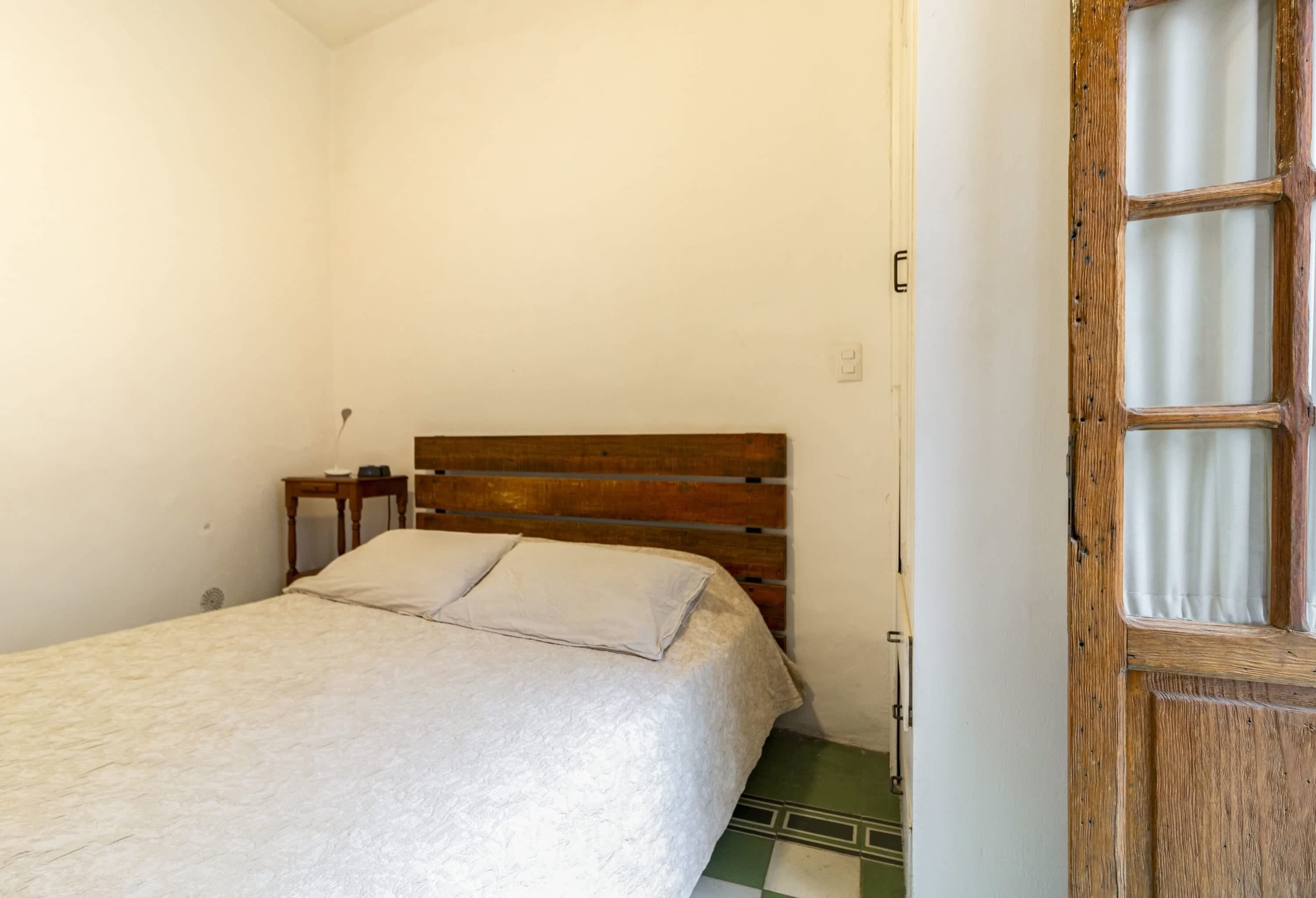 A bedroom with a wooden headboard, a bed with a white bedspread, a bedside table, and a door with a window.