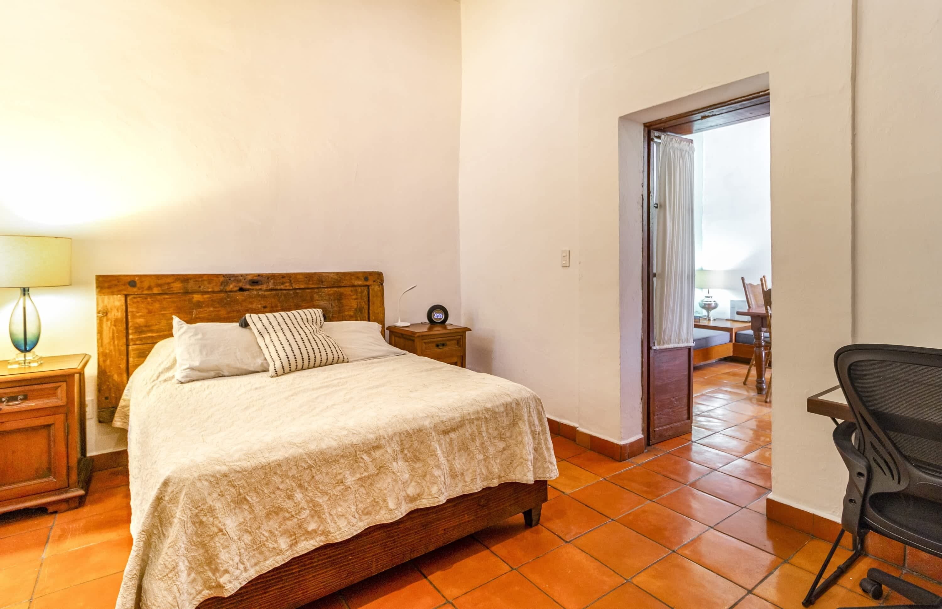 A bedroom with a wooden bed, a nightstand with a lamp, a chair, and a view of a balcony through an open door.