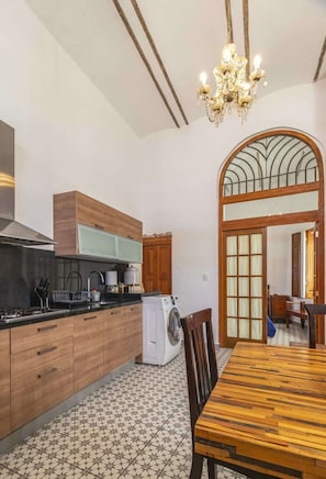 A modern kitchen with a washing machine, wooden cabinets, a chandelier, and a dining area with a patterned floor.