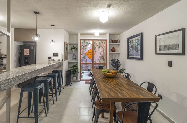 A modern kitchen with a granite countertop, a wooden dining table, and bar stools. There are two pendant lights, a refrigerator, and a fan on the wall.