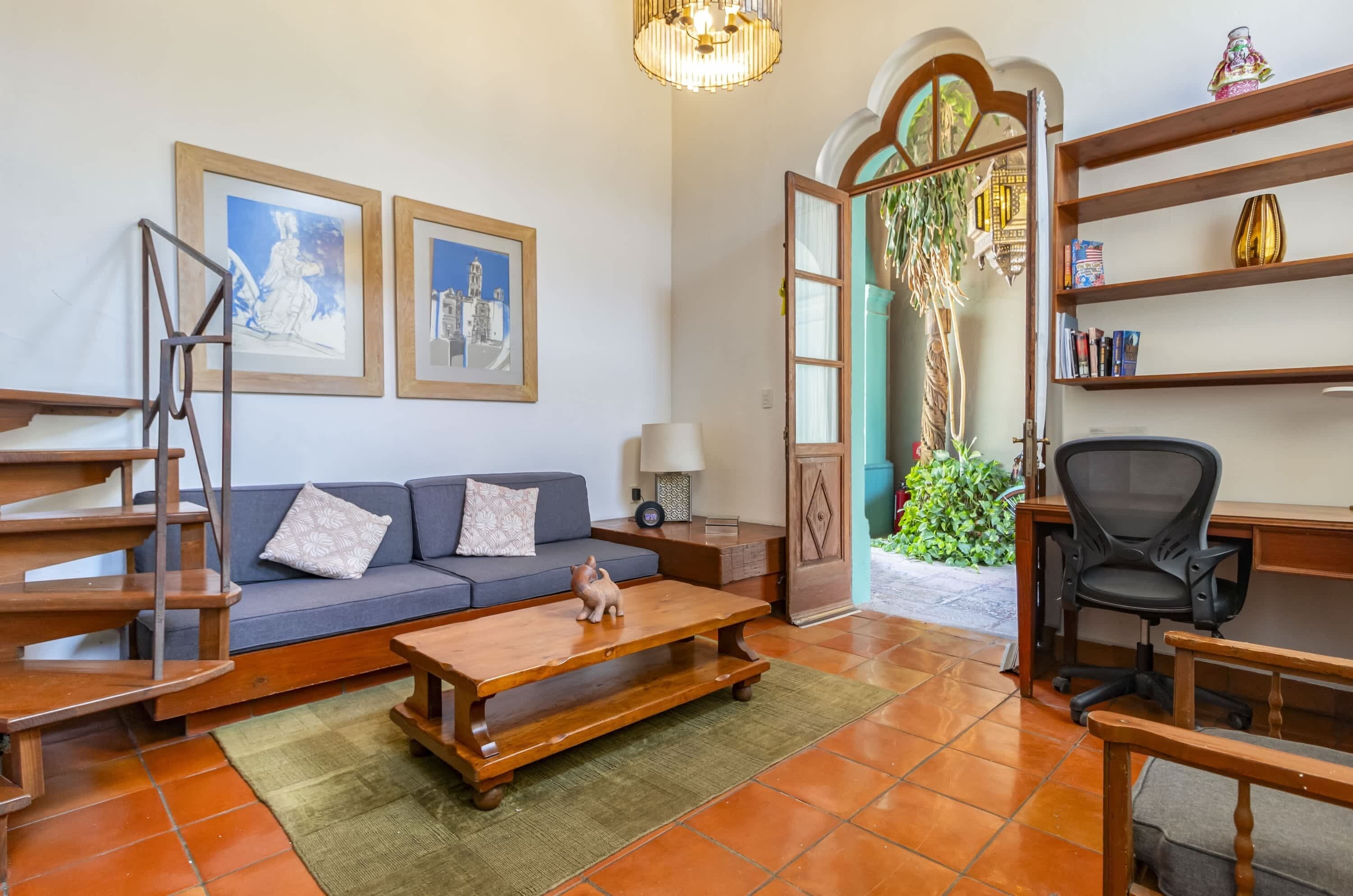 A living room with a sofa, coffee table, and a staircase. There are framed pictures on the wall and a desk with a chair.