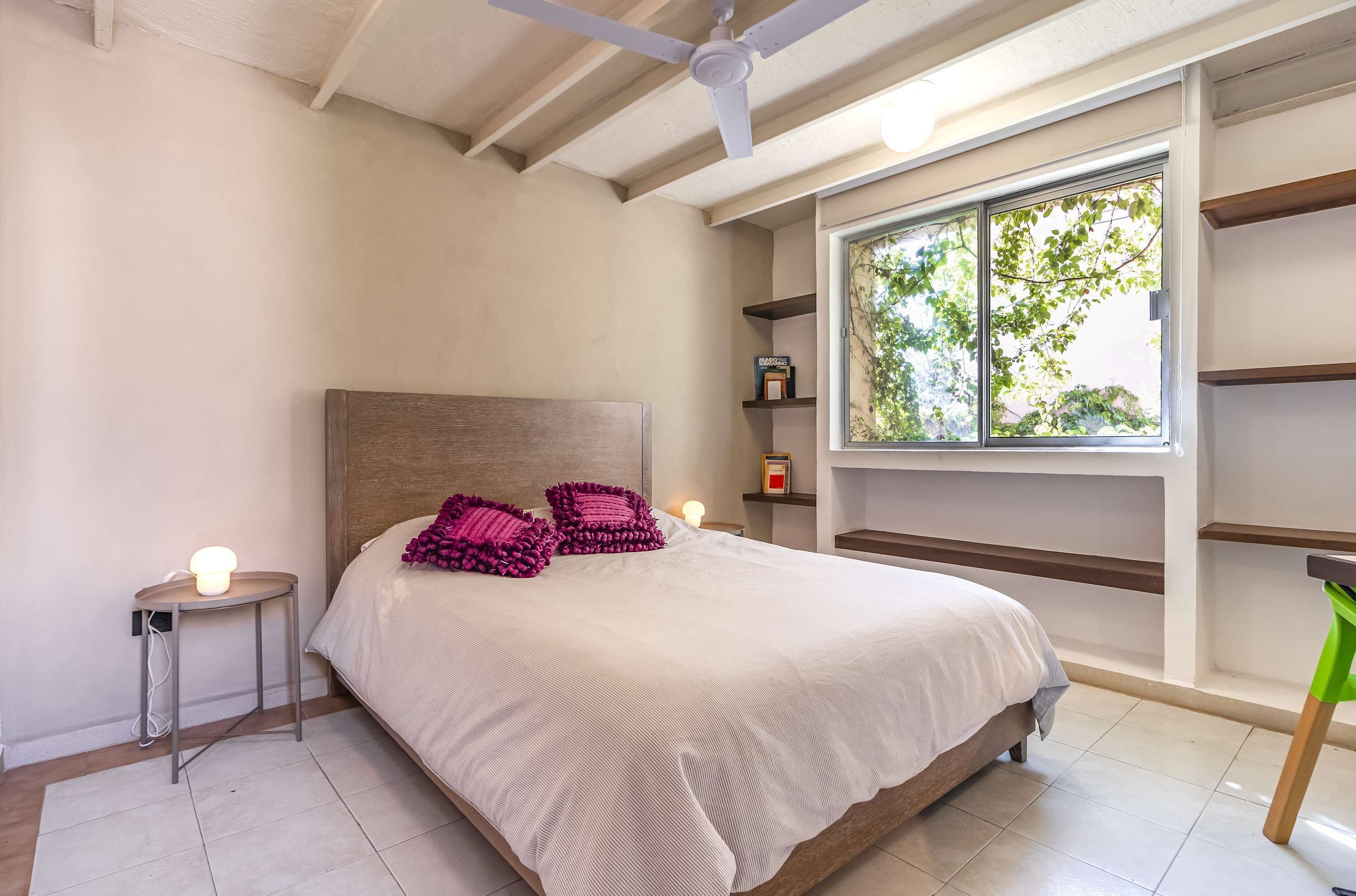 A bedroom with a bed, a ceiling fan, a window with a view of greenery, and a bedside table with a lamp.