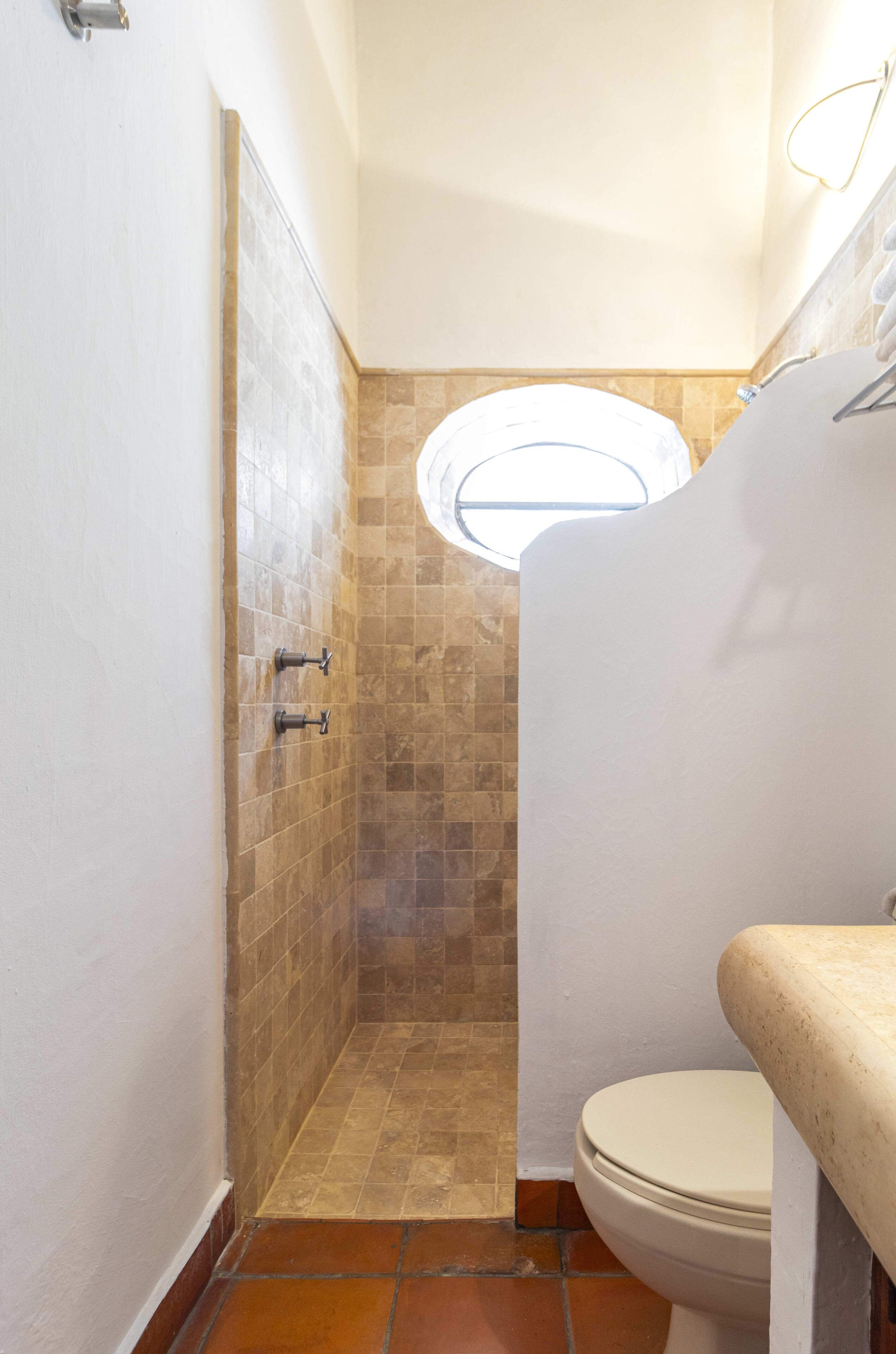 Une salle de bain avec des toilettes blanches, une douche aux carreaux beiges et une petite lucarne.