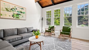 A modern living room with a grey sofa, two green armchairs, a round coffee table with a vase of flowers, and large windows with a view of greenery. - Premier 3 BD with Pool, Outdoor Dining (Santa Rosa)