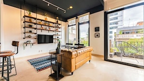 A modern living room with a brown leather sofa, a coffee table, and a TV mounted on the wall. There are shelves with plants and decorative items, and a large window providing a view of the city. - NEW Perfect Location with Free Parking (Mexico City)