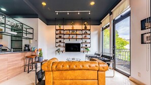 A modern living room with a leather sofa, a coffee table, and a TV mounted on the wall. There is a kitchen area with bar stools and a dining table. The room has large windows with a view of the outside. - NEW Perfect Location with Free Parking (Mexico City)