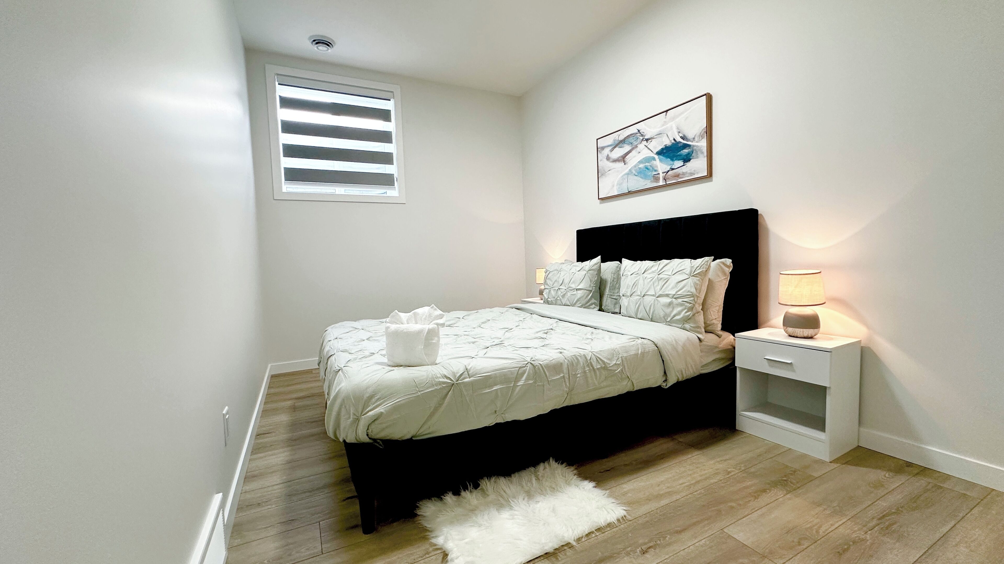 Een slaapkamer met een bed, een nachtkastje, een lamp, een wit vloerkleed, een raam met zonwering en een wanddecoratie.