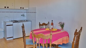 A small kitchen with a gas stove, a sink, and a white refrigerator. There is a dining table with a pink tablecloth and two wooden chairs. A wine bottle and glasses are on the table. - Apartments Jug - Comfort Studio Apartment with Balcony (Lopar)