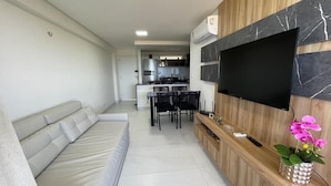 A modern living room with a sofa, a TV mounted on a wooden wall, a dining area with chairs, and a kitchen in the background. - Pé na Areia em Ponta de Campina, Paraíba (Cabedelo)