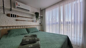 Makuuhuoneessa on vihreä, kuviollinen peitto, puinen pääty ja valkoinen tiiliseinä, johon on asennettu ilmastointilaite.