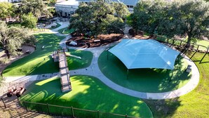 An aerial view of a park with a large tent, green lawns, a playground, and a building with a blue roof. - HOT TUB Tranquil Escape Pool Gym (Round Rock)