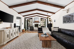A spacious living room with a high ceiling, exposed wooden beams, a fireplace, a black leather sofa, and a patterned rug. - HOT TUB Tranquil Escape Pool Gym (Round Rock)