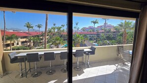 A balcony with four bar stools and a table, overlooking a resort area with palm trees and buildings with red roofs.
