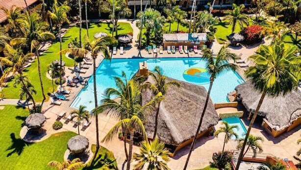 An aerial view of a resort with a central swimming pool, surrounded by palm trees and thatched-roof structures.