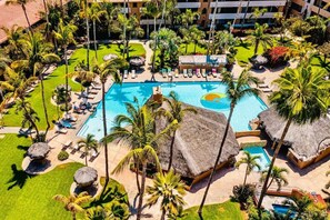 Oversigtsbillede af et resort med en central swimmingpool, omgivet af palmer og stråtækte bygninger.