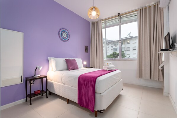 A bedroom with a bed, a bedside table, a lamp, a window with curtains, and a view of buildings outside.