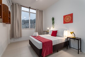 A hotel room with a bed, a bedside table with a lamp, a window with curtains, a wall-mounted shelf, and a decorative wall piece.