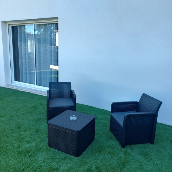 Un patio con dos sillas de mimbre negro y una mesa cuadrada a juego, frente a una pared blanca y un ventanal grande.