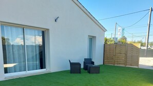 Un edificio moderno con un gran ventanal corredero, un pequeño patio con mobiliario exterior y un césped bien cuidado.