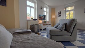 Ein Schlafzimmer mit einem Bett, einem grauen Sessel, einem Heizkörper, einem Fernseher und einem Fenster mit Blick auf Bäume.