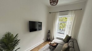 Ein Wohnzimmer mit an der Wand befestigtem Flachbildfernseher, einer Couch mit Kissen, einem kleinen Beistelltisch, einer Topfpflanze und einem Fenster mit transparenten Vorhängen.