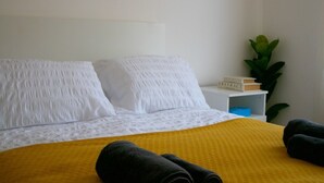 Ein ordentlich bezogenes Bett mit weißen Leinen und einer gelben Decke, ein kleiner Nachttisch mit einem Buch und eine grüne Pflanze.