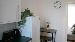 Eine kompakte Küche mit weißen Schränken, einem weißen Kühlschrank, einer Wanduhr und einem kleinen Tisch mit einer Pflanze.