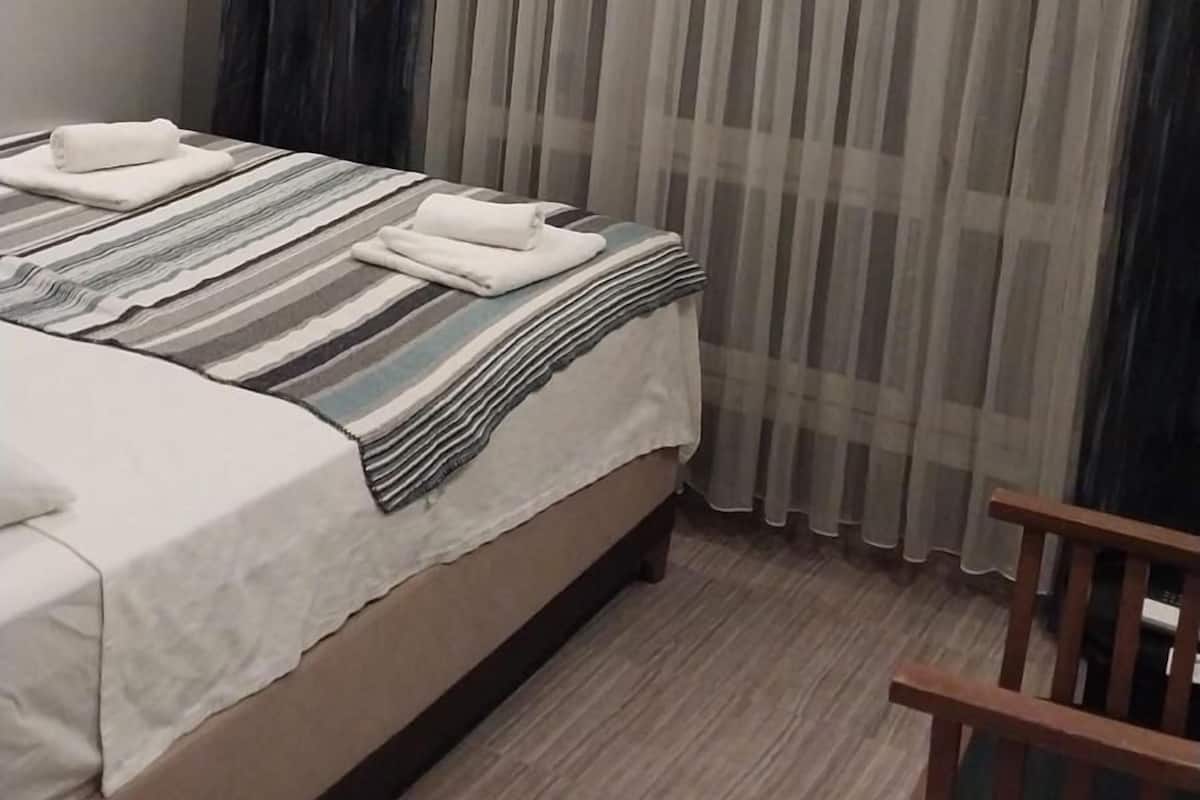 Hotellihuone, jossa on sänky, puinen tuoli ja ikkuna, jossa on ohuet verhot.