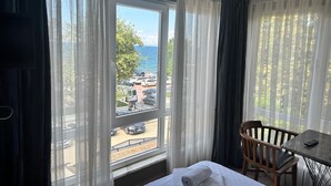 Een hotelkamer met een bed, een klein tafeltje en een stoel. Er is een groot raam met dunne gordijnen en uitzicht op een parkeerplaats en bomen.