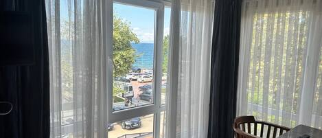 Habitación de hotel con cama, una mesita y una silla. Hay un ventanal con cortinas ligeras y vistas a un estacionamiento y árboles.