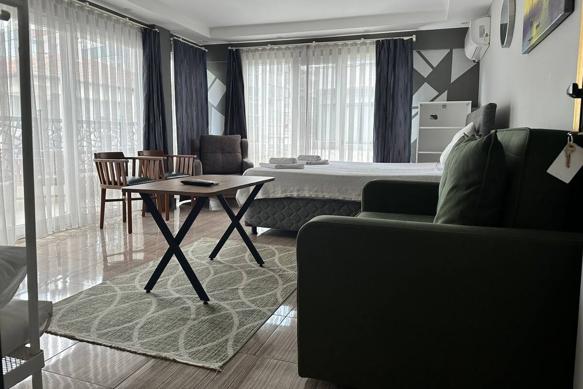 Moderni hotellihuone, jossa on sänky, vihreä nojatuoli, puinen ruokapöytä ja sohva.